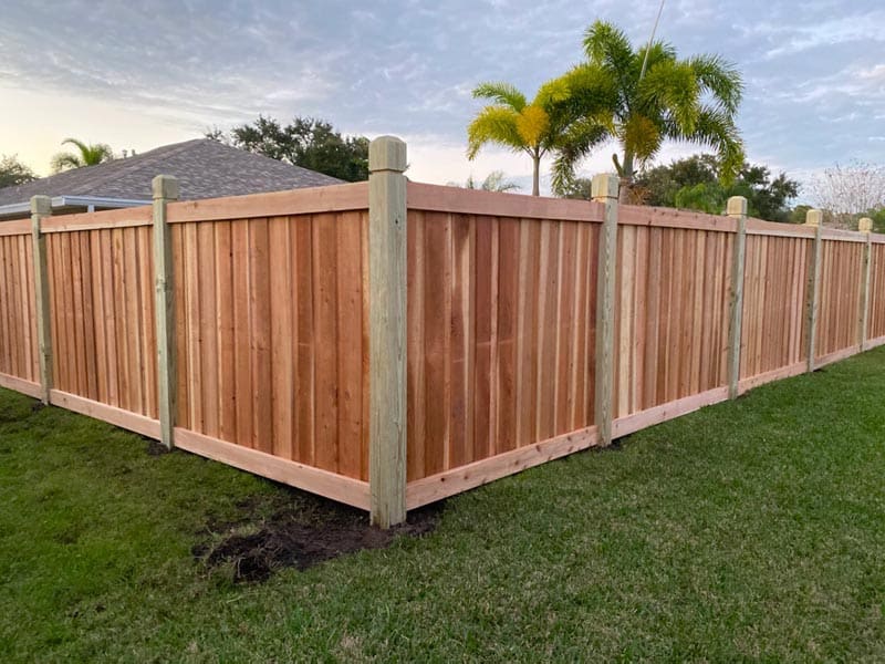 A corner of a wooden fence made of vertical planks with sturdy posts stands on a grassy lawn. In the background, there are palm trees and a house visible under a partly cloudy sky.