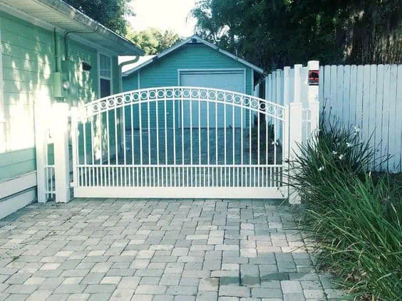 A white picket fence gate opens to a driveway leading to a light green garage. The paved path is surrounded by greenery and a light green house is visible on the left. .