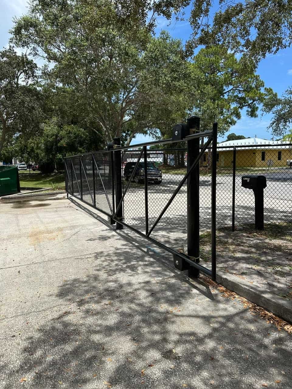 A black metal sliding gate is partially open on a sunny day. The gate is set within a chain-link fence. There are trees and a yellow building in the background, and a vehicle is visible beyond the gate. Shadows from the trees cover part of the ground.