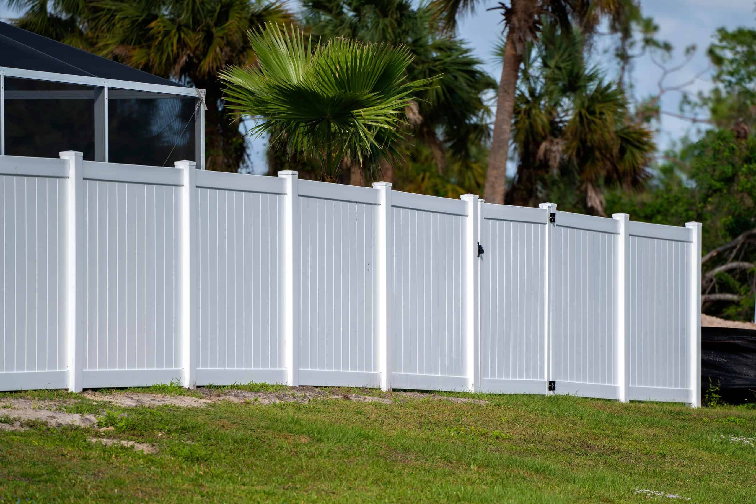 A white vinyl privacy fence surrounds a grassy yard with palm trees in the background. The fence has evenly spaced posts and a slight upward slope, contrasting with the lush, green landscape and clear blue sky above.