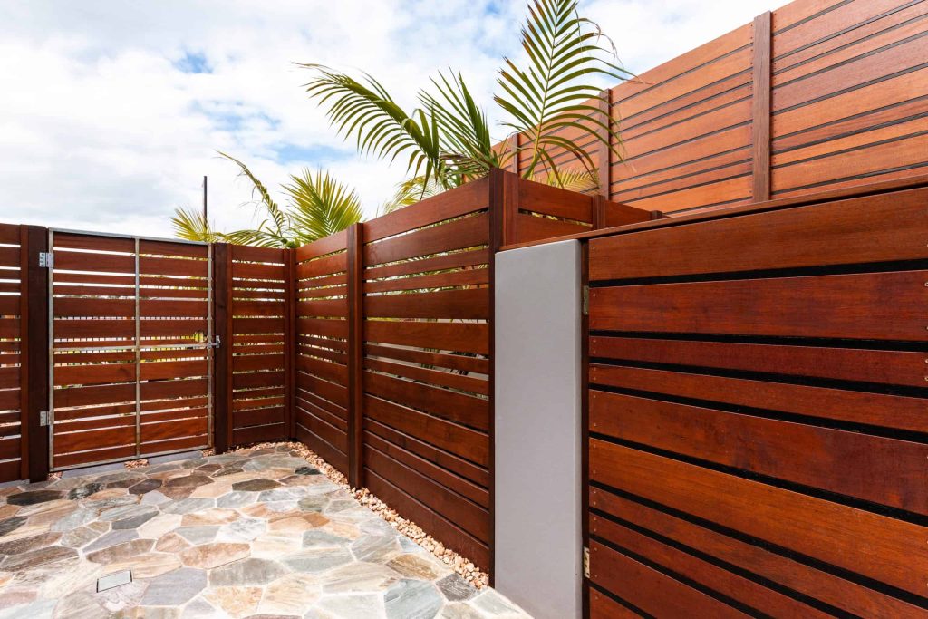 Wooden privacy fence with horizontal slats, featuring a seamless wooden gate installation, encloses a patio area with stone paving. Tropical plants peek from behind as the partly cloudy sky provides a bright and natural backdrop.