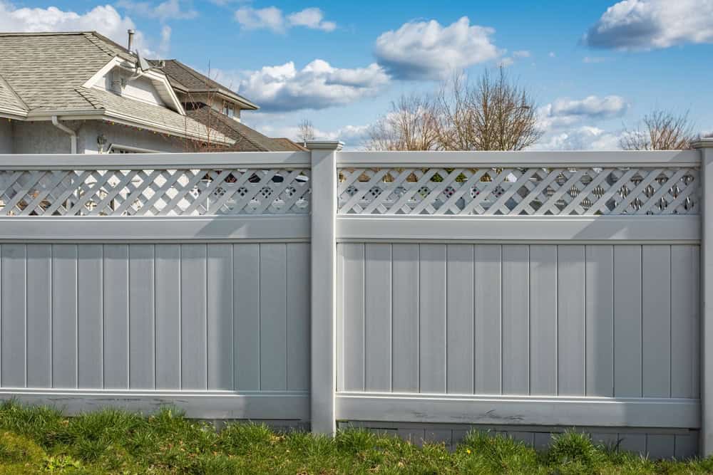 A white vinyl fence with lattice detailing on top stands in a green grassy yard. In the background, a residential house is partially visible under a blue sky with scattered clouds.