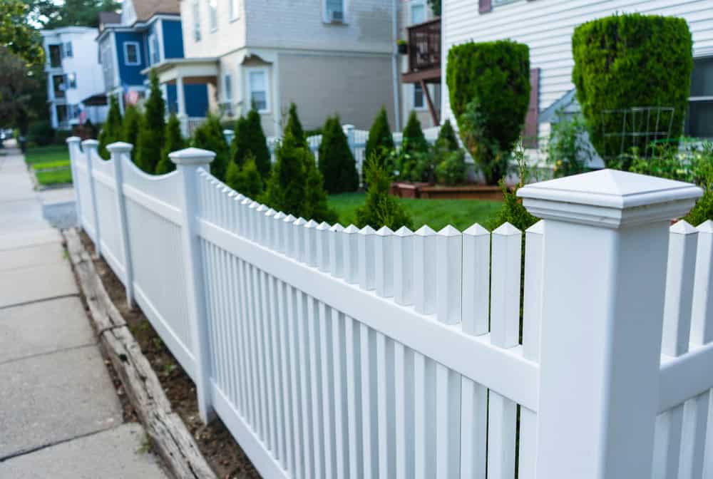 A white picket fence lines a sidewalk, with neatly trimmed evergreen shrubs behind it. In the background, there are blue and white residential houses with balconies and stairs. The scene is well-lit, suggesting a sunny day.