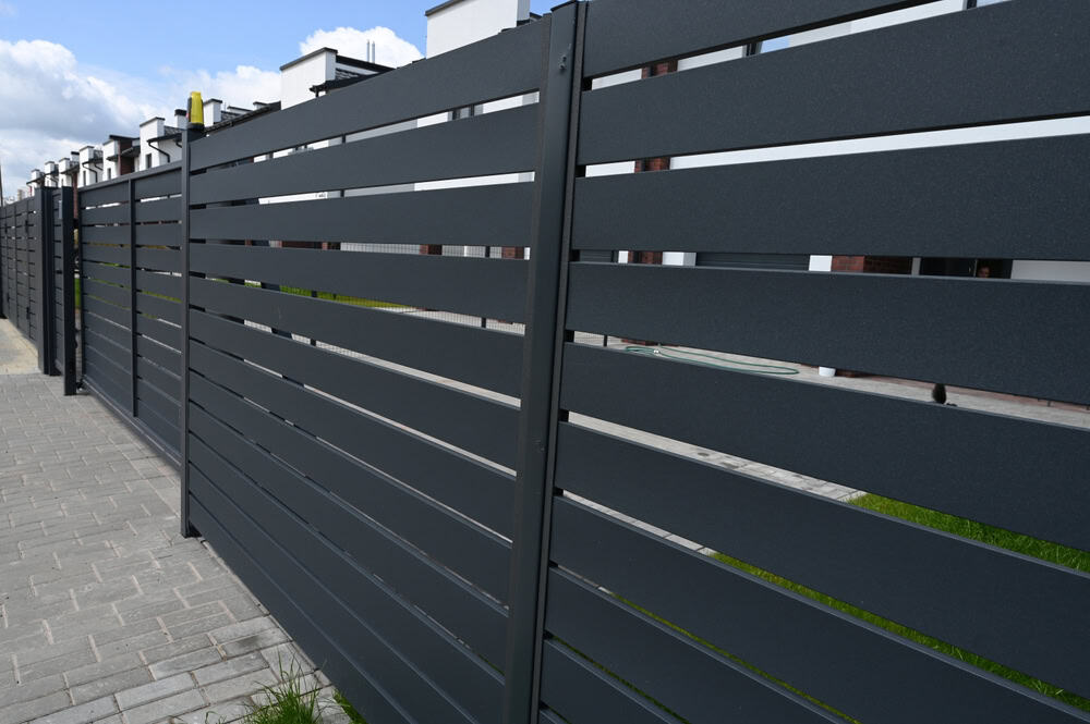 A modern, dark gray metal fence with horizontal slats lines a paved walkway. Behind the fence are buildings with white walls and windows, partially visible against a backdrop of a cloudy blue sky.