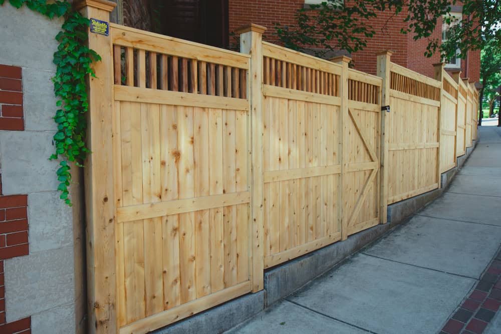 A wooden fence runs alongside a sidewalk bordered by a brick building. The fence has vertical slats at the top and a closed gate. Lush green vines climb up part of the building near the fence.