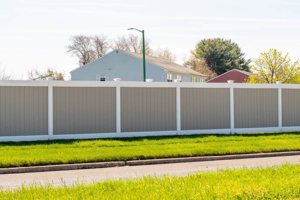 A suburban neighborhood scene with a tall gray privacy fence with white trim running along a green lawn. In the background, there are houses partially visible behind the fence, and trees with sparse leaves. A street lamp stands near the fence.