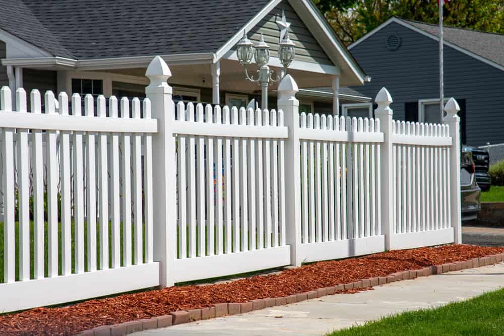 A white picket fence lines the front yard of a suburban house. The fence is complemented by red mulch along its base. The house is gray with a peaked roof and a porch. Another house is partially visible in the background.