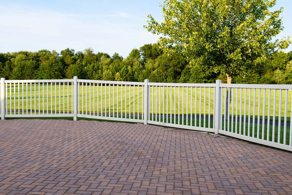 A white picket fence curves around a stone-paved patio, enclosing a grassy lawn with a tree. In the background, a line of lush green trees stretches across the horizon under a clear blue sky.