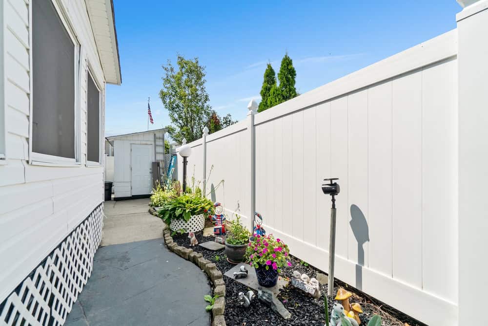 A narrow backyard pathway with a white fence on one side and a house on the other. The path is decorated with flower pots, garden gnomes, and solar lights. A small shed and a tree are visible in the background. The sky is clear and blue.