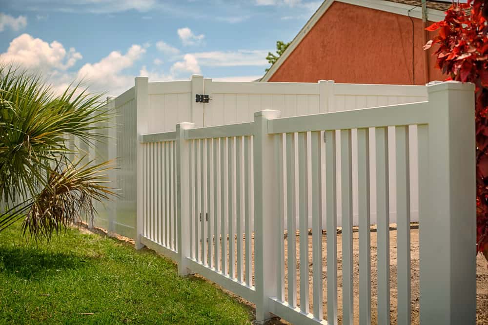 A white fence with vertical slats borders a green lawn, extending toward a pink house in the background. Red foliage is visible on the right, and the sky is clear with a few clouds.