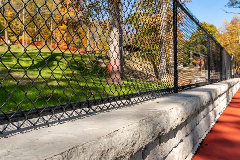 A chain-link fence atop a stone wall surrounds a grass field with autumn trees. A red running track runs alongside the fence. .