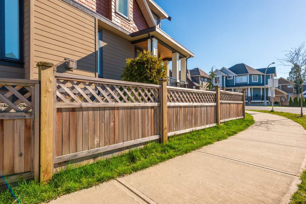 A suburban street view featuring a wooden fence with lattice detailing in front of a modern house. The sidewalk curves alongside neatly trimmed grass, while similar houses are visible in the background under a clear blue sky.