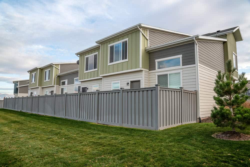 A row of modern townhouses with green and gray siding, large windows, and an adjoining fenced backyard. The sky is partly cloudy, and the lawn is neatly trimmed.