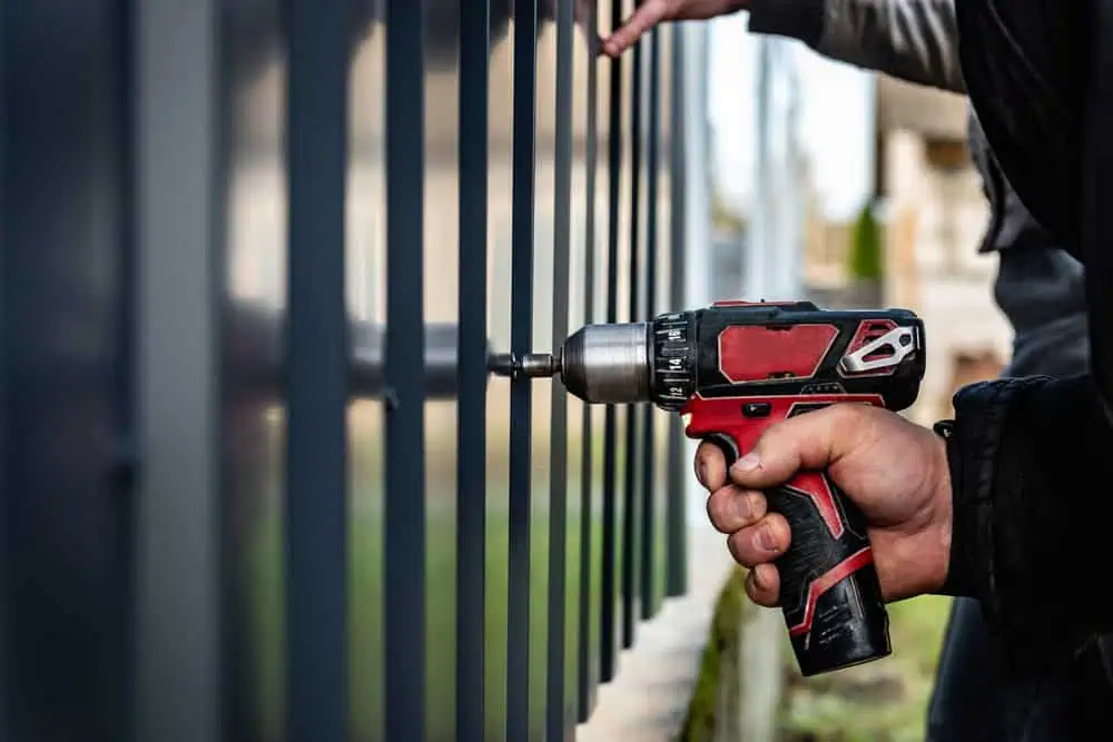 Close-up of a person using a red and black cordless drill to fasten a bolt on a black metal fence. The background is blurred, focusing on the hand holding the drill and the fence.