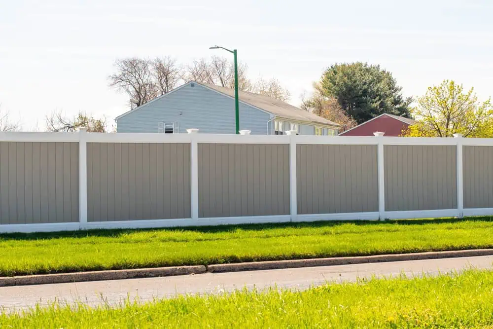 A suburban neighborhood scene with a tall gray privacy fence with white trim running along a green lawn. In the background, there are houses partially visible behind the fence, and trees with sparse leaves. A street lamp stands near the fence.