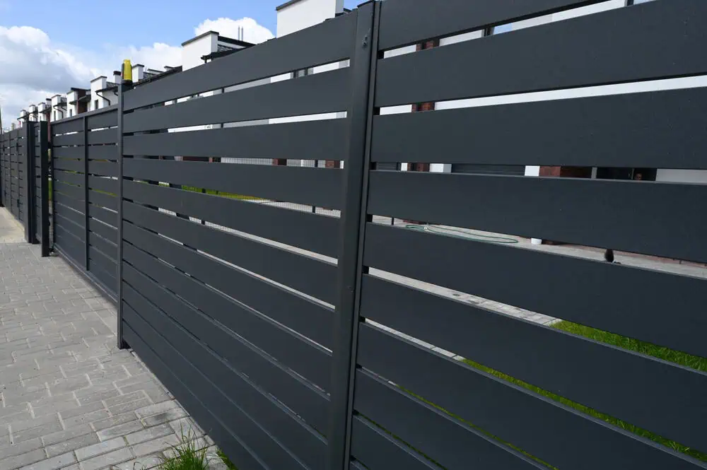 A modern, dark gray metal fence with horizontal slats lines a paved walkway. Behind the fence are buildings with white walls and windows, partially visible against a backdrop of a cloudy blue sky.
