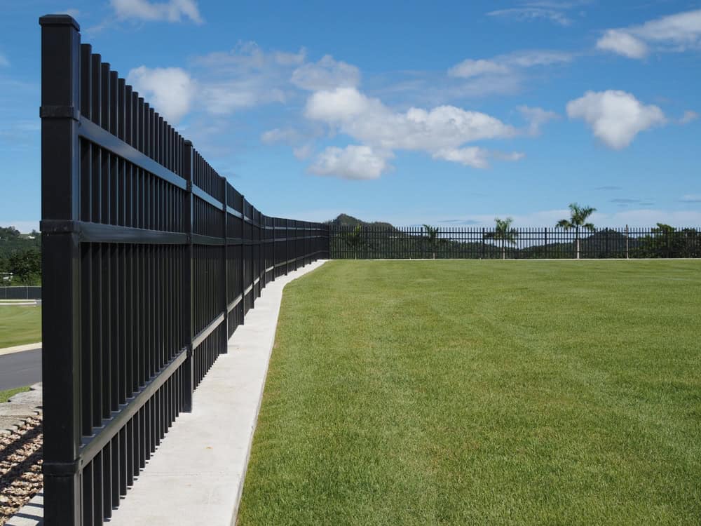 A long, black metal fence runs along a neatly trimmed green lawn under a blue sky with scattered clouds. A distant hill and a few trees are visible in the background.