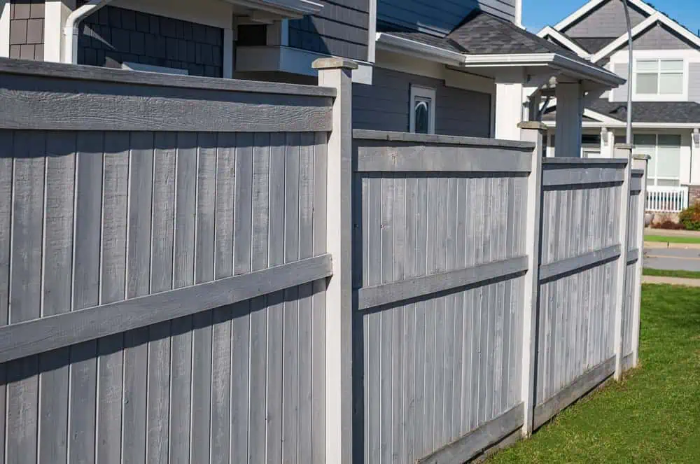 A view of a wooden fence lining the side of a modern suburban house. The fence is painted gray, and the house features gray siding with white trim. Bright sunlight casts shadows, and a manicured lawn is visible alongside the house.