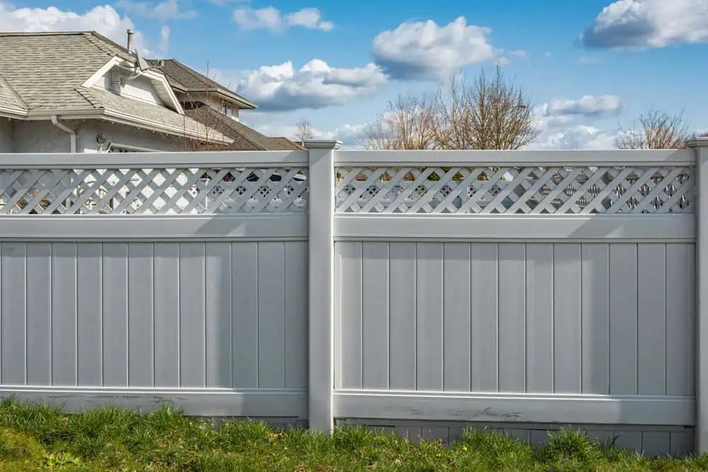 A white vinyl fence with lattice detailing on top stands in a green grassy yard. In the background, a residential house is partially visible under a blue sky with scattered clouds.