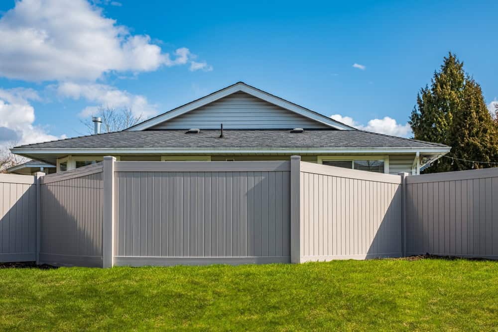 A house with a gray roof is partially visible behind a tall, light gray privacy fence. There's a bright green lawn in the foreground and a blue sky with scattered clouds in the background.