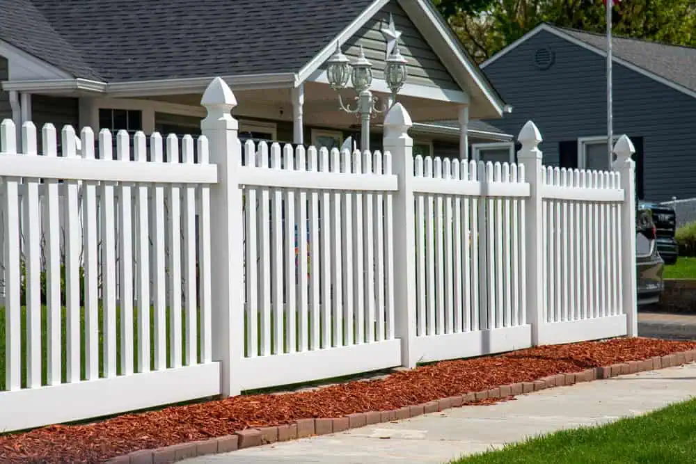 A white picket fence lines the front yard of a suburban house. The fence is complemented by red mulch along its base. The house is gray with a peaked roof and a porch. Another house is partially visible in the background.