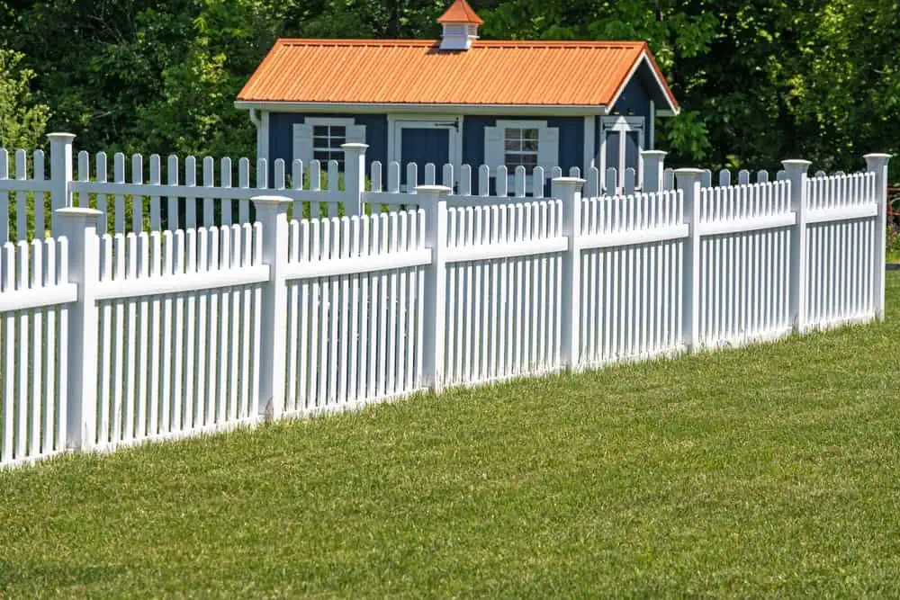 A white picket fence runs diagonally across a green lawn, leading to a small blue shed with an orange roof, set against a backdrop of leafy green trees.