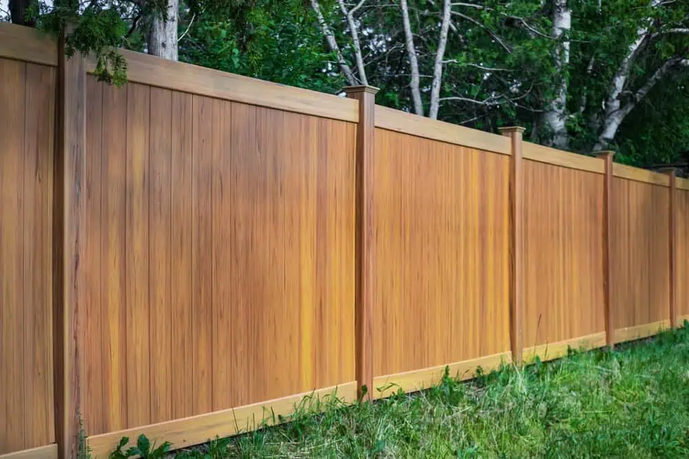 A wooden privacy fence stands in a lush, green yard. The fence has evenly spaced vertical slats and horizontal beams, with posts extending above. Dense foliage and trees are visible behind the fence, creating a serene, natural backdrop.