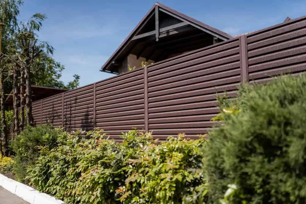 A brown wooden fence partially obscures a house with a dark roof in the background. In the foreground, there are green shrubs and trees under a clear blue sky.