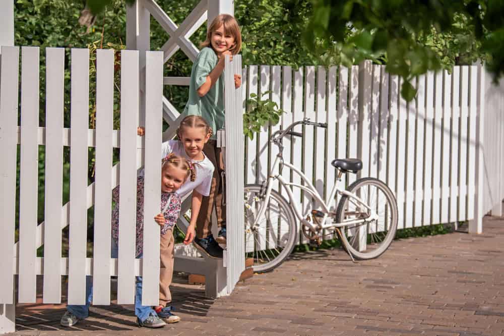 Three children peek playfully through a white picket fence, with a white bicycle parked nearby. The ground is paved, and lush greenery is visible in the background.