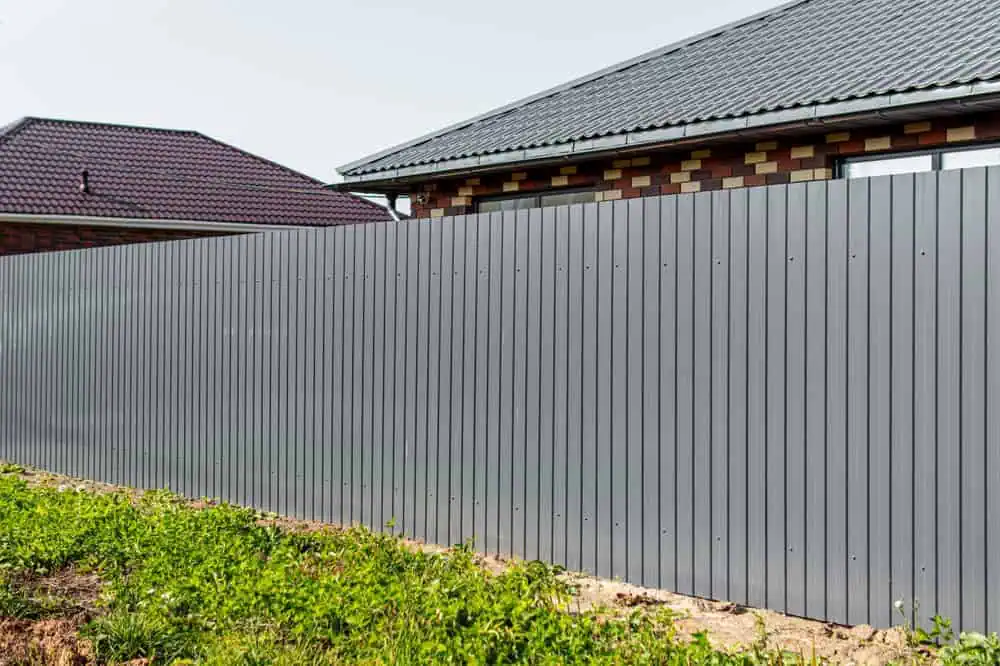 A long, gray metal fence borders a property with two brick houses in the background. The fence is adjacent to a patch of green grass. The sky is clear.