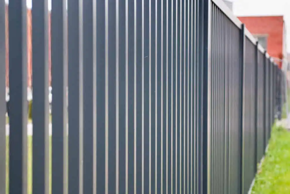 Close-up of a modern black metal fence with vertical bars, set against a blurred background of grass and red brick buildings. The fence is viewed at an angle that emphasizes its sleek design and shadows.