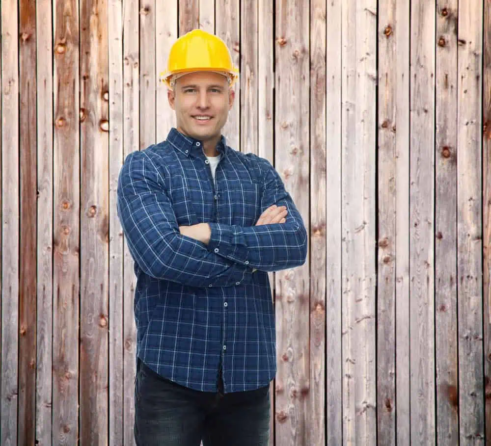A man wearing a yellow hard hat and a blue plaid shirt stands with his arms crossed in front of a wooden fence. He is smiling and appears confident.